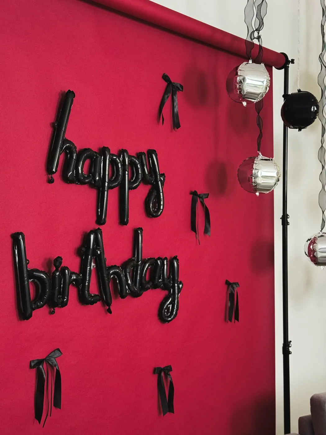 Tumši sarkans fons ar melnu balonu uzrakstu „happy birthday” un melniem lentes lociņiem