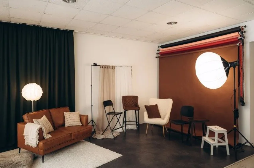 Fotostudija: brūns papīra fons, liels apaļš softbokss, konjaka krāsas ādas dīvāns, balts krēsls un tumši aizkari